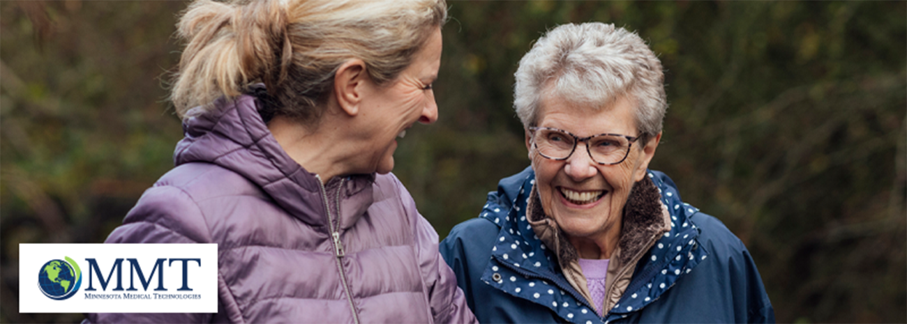 Image shows older woman and female caregiver laughing together in outdoor environment